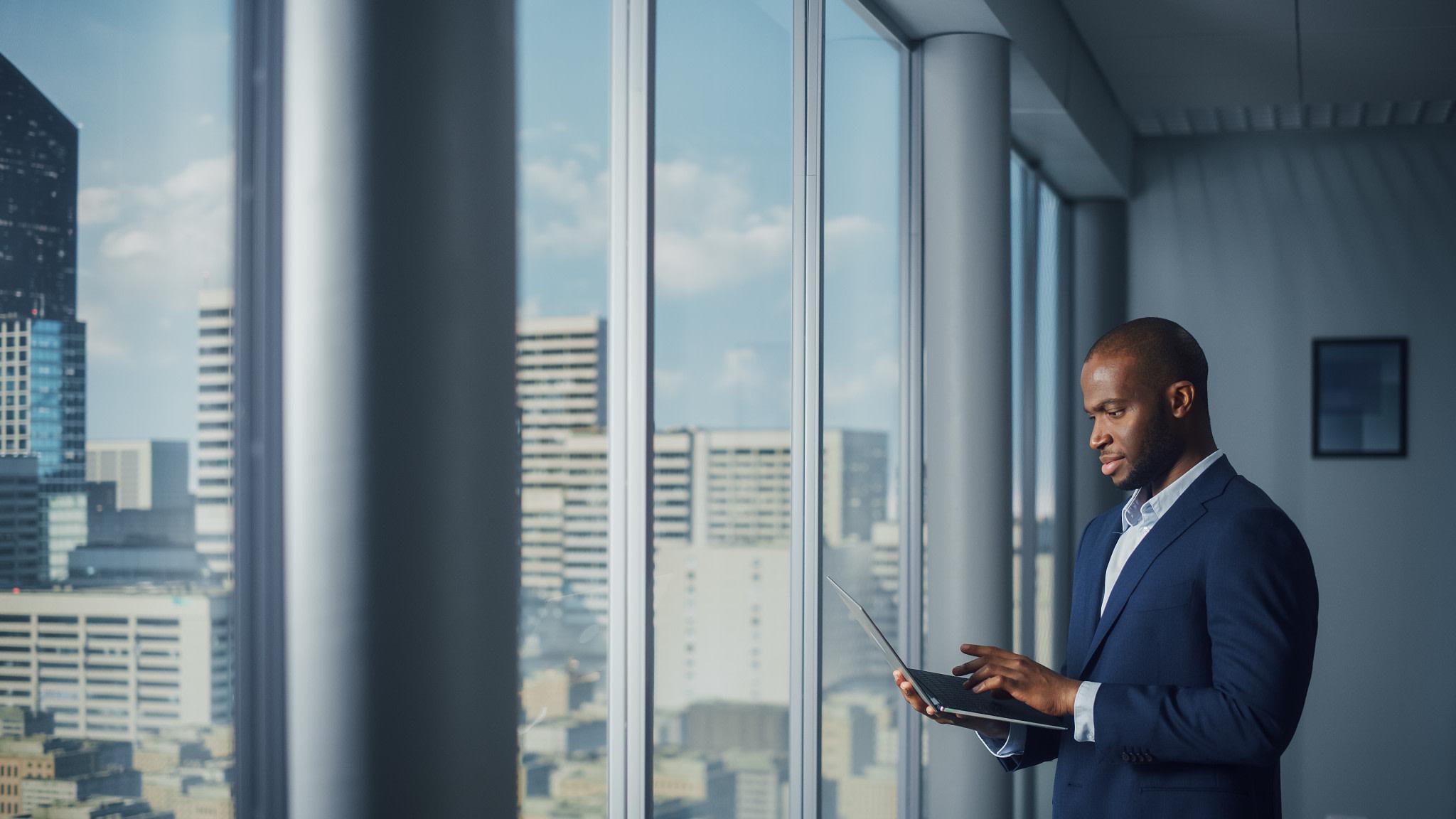 Businessman standing by office window with a laptop.